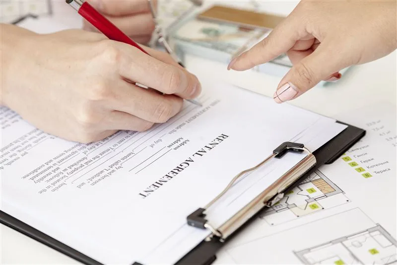 One hand signing a document on a clipboard while another hand points to the page